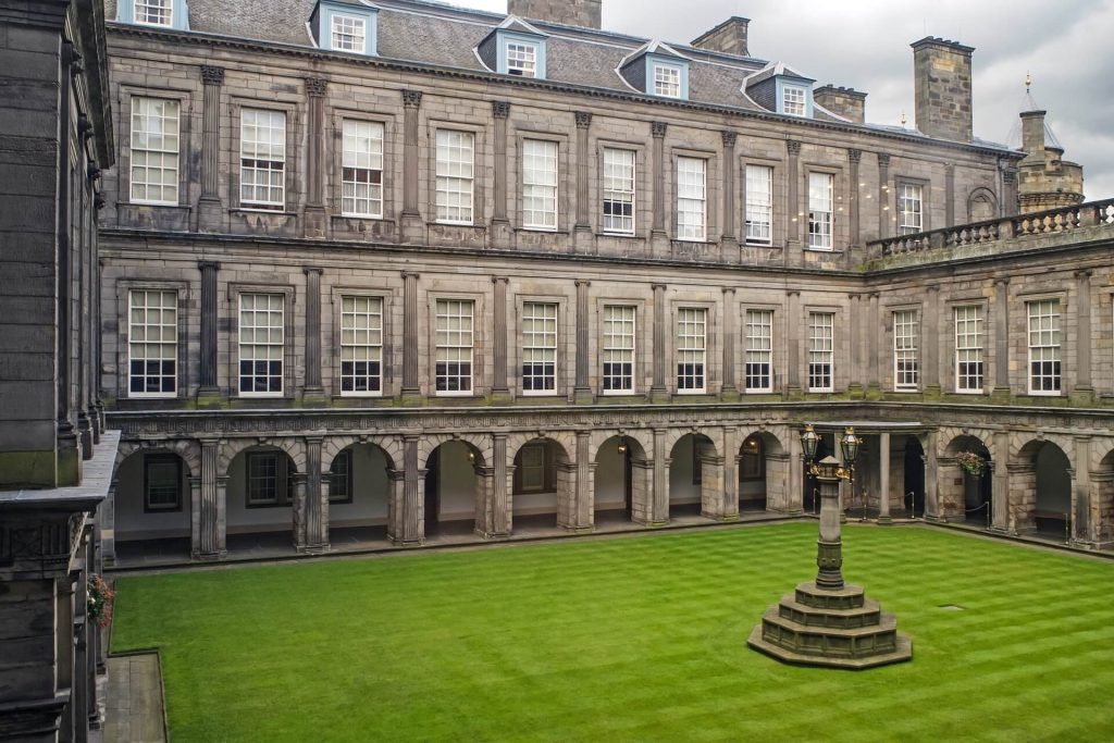 A large, grey palatial building and green lawn with a statue in the centre